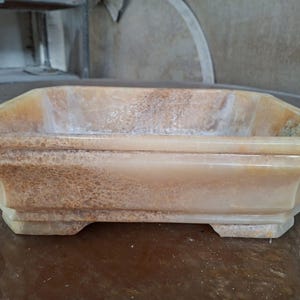 May include: A rectangular, light-colored stone planter with an octagonal shape. The planter has a textured surface with brown and white veining. The edges are slightly flared, and the base has small feet. The planter is empty.