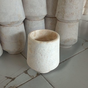 May include: A collection of off-white stone cups featuring a textured, ribbed exterior surface. Each cup has a tapered cylindrical shape with a smooth, polished interior rim. The stone displays natural beige and cream marbling throughout the surface.
