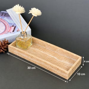 May include: A rectangular beige stone tray with a textured surface, measuring 30 cm in length, 10 cm in width, and 4 cm in height. A clear glass square bottle containing a golden liquid and two white flower-shaped reed diffusers sits inside.