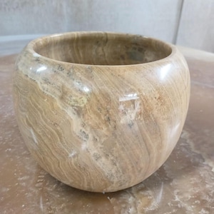 Könnte beinhalten: Eine runde, beige Steinschale mit polierter Oberfläche. Die Schale hat eine weite Öffnung und eine abgerundete Form. Der Stein weist natürliche Muster und Farbvariationen mit braunen und grauen Streifen auf.