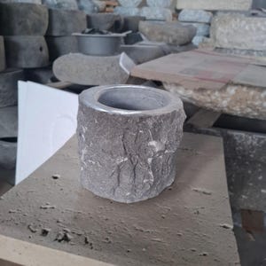 Peut inclure: Un bougeoir en pierre grise avec un extérieur rugueux et texturé et un bord lisse de couleur argentée. Le support cylindrique repose sur une surface en pierre, avec d'autres objets en pierre en arrière-plan. Le bougeoir mesure environ 10 cm de haut.