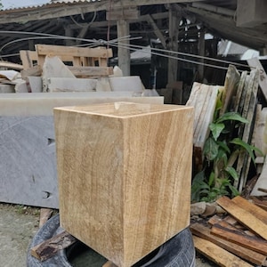 May include: A large, square, light brown stone planter with a textured surface. The planter is sitting on a black tire. The background includes a wooden structure and other stone slabs.