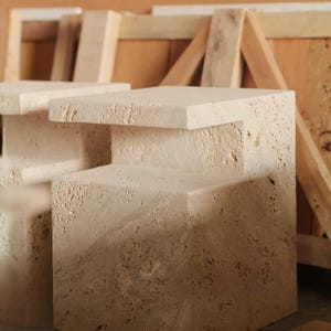 May include: A beige travertine side table featuring a stepped, geometric silhouette with a porous, natural stone texture. The piece has a flat top surface and a lower shelf section, constructed from solid, light-coloured stone with visible pits.