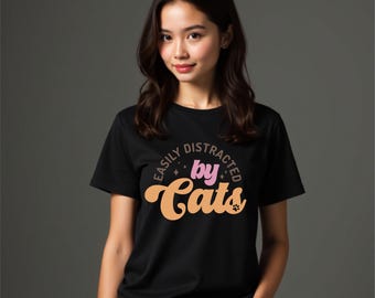 Puede incluir: Camiseta negra con el texto "Easily Distracted by Cats" en una fuente retro. El texto es en tonos rosa y naranja. La camiseta tiene cuello redondo y mangas cortas.