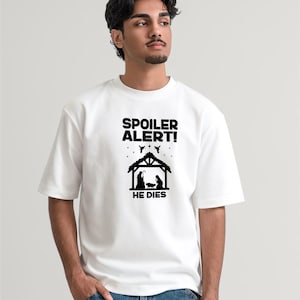 May include: White t-shirt with the text "SPOILER ALERT!" above a black silhouette of a nativity scene and the words "HE DIES". The shirt is being worn by a person in blue jeans.