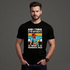 Può includere: T-shirt nero con grafica di un saldatore e la scritta "AND I THINK TO MYSELF WHAT A WONDERFUL WELD". La grafica mostra un saldatore con casco rosso e giacca blu, con scintille gialle.