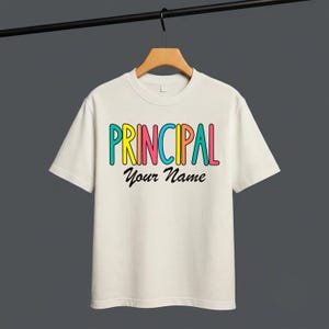 Op de afbeelding: Een crèmekleurig t-shirt met korte mouwen en het woord PRINCIPAL in kleurrijke, afgeronde blokletters in turquoise, geel, roze en oranje. Daaronder staat de tekst Your Name gedrukt in een zwart, cursief, handgeschreven lettertype.