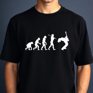 Może przedstawiać: Czarny t-shirt z białym nadrukiem przedstawiającym ewolucję człowieka, zakończoną postacią grającą na gitarze. Projekt w humorystyczny sposób ilustruje ewolucję człowieka w gitarzystę, zabawne spojrzenie na muzykę.
