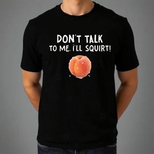 May include: A black short-sleeved t-shirt featuring the white text DON'T TALK TO ME, I'LL SQUIRT! above a graphic of a single peach with water droplets on its skin. The design is centered on the chest of the garment.