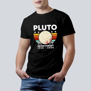 Può includere: T-shirt nera con un design grafico. Il design presenta la parola "PLUTO" sopra un'immagine stilizzata di Plutone. Sotto l'immagine del pianeta, il testo recita "NEVER FORGET 1930 - 2006". Il design include stelle e strisce colorate.