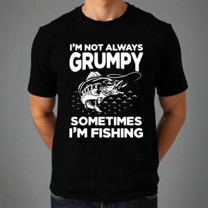 Könnte beinhalten: Schwarzes T-Shirt mit dem weißen Text "I'M NOT ALWAYS GRUMPY SOMETIMES I'M FISHING". Eine detaillierte Fischillustration mit Angelhaken befindet sich unter dem Wort "GRUMPY". Das Shirt besteht aus einem weichen Material.