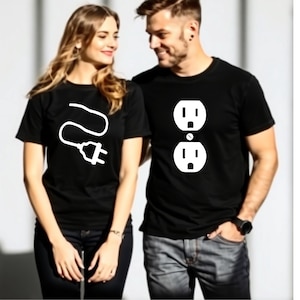 Pode incluir: Duas pessoas vestindo camisetas pretas combinando. Uma camisa apresenta um gráfico de plugue branco, a outra um gráfico de tomada branco. Ambas são de manga curta e provavelmente de algodão. O casal sorri um para o outro, sugerindo um tema de relacionamento.