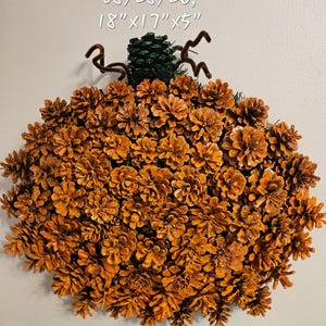 May include: A pumpkin-shaped wreath crafted from numerous brown pinecones, featuring a dark green pinecone stem and brown wire tendrils. The text 08/28/26, 18"x17"x5" is printed above the wreath, indicating the date and dimensions.