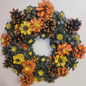 May include: A circular wreath featuring painted pinecones and flower-shaped pinecone arrangements in shades of orange, yellow, black, and dark green. The text 09/08/26, 13" is visible at the top, indicating a diameter of 13 inches.