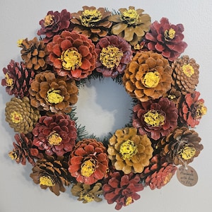 May include: A circular wreath constructed from numerous pinecones painted in shades of red, brown, and yellow. The pinecones are arranged to resemble flowers with bright yellow centers. A small circular tag reads Handmade with love by Lily.