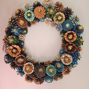 May include: A circular wreath constructed from numerous pinecones in various sizes. The pinecones are painted in shades of blue, green, orange, brown, and white, with many featuring gold-colored centers that resemble flower petals.