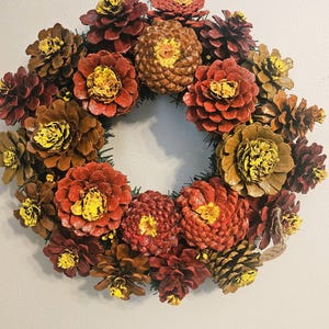 May include: A circular wreath constructed from numerous pinecones, with many individual pinecone scales painted in shades of red, orange, and brown, and the centers of the pinecone clusters accented with bright yellow paint.