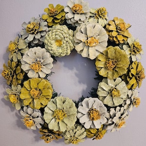 May include: A decorative wreath made of painted pinecones in shades of yellow, cream, and green. The pinecones are arranged in a circular pattern, resembling flowers. The wreath is a natural, rustic home decor item.