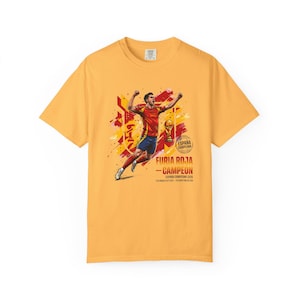 May include: A mustard yellow t-shirt featuring a graphic of a soccer player celebrating a victory. The design includes the text "FURIA ROJA - CAMPEON", "ESPAÑA CAMPEONA", and "FIFA WORLD CUP 2010 - CELEBRATION EDITION" with a golden trophy illustration.