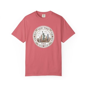 May include: A coral-colored short-sleeve t-shirt featuring a circular graphic on the chest. The design displays the Statue of Liberty and the New York City skyline, surrounded by a floral wreath, with the text NEW YORK and USA printed in black.