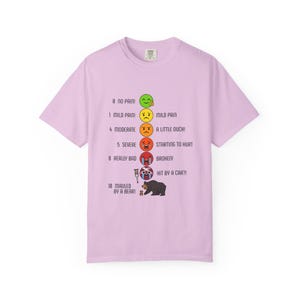 Peut inclure: T-shirt lilas avec un graphique d'échelle de douleur. L'échelle va de "0 Pas de douleur" à "10 Attaqué par un ours !" avec des émoticônes et des descriptions telles que "Douleur légère" et "Heurté par une voiture ?!".