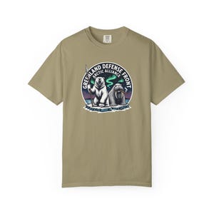 Puede incluir: Camiseta color arena con un gráfico circular que representa un oso polar con una lanza y una morsa. El gráfico dice "Greenland Defense Front Arctic Alliance". El diseño incluye una aurora boreal y témpanos de hielo.