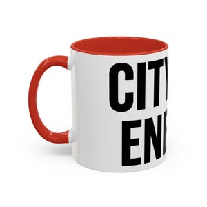 Peut inclure: Mug en céramique blanche avec intérieur et anse rouges. Le mug porte les mots "CITY" et "EN" en grandes lettres majuscules noires. Le mug est conçu pour les boissons chaudes ou froides.
