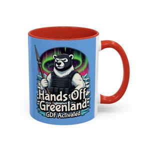 Puede incluir: Taza de cerámica blanca con interior y asa rojos. La taza presenta un oso de dibujos animados con chaleco táctico y lanza, con el texto "Hands Off Greenland" y "GDF Activated" sobre fondo azul.