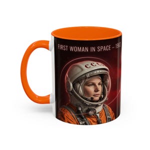 Könnte beinhalten: Ein Keramikbecher mit leuchtend orangefarbenem Henkel und Innenraum. Die Außenseite zeigt eine Grafik eines Astronauten mit weißem Helm und der Aufschrift CCCP vor einem dunkelroten Weltraumhintergrund mit dem Text FIRST WOMAN IN SPACE - 1963.