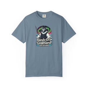 Puede incluir: Camiseta azul de manga corta con un gráfico de un oso sosteniendo una lanza, sobre un fondo de aurora boreal. El texto "Hands Off Greenland" y "GDF Activated" están debajo de la ilustración del oso.