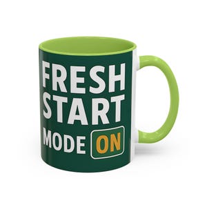 May include: A white ceramic mug with a green interior and handle. The mug features the text "FRESH START MODE ON" in white block letters against a dark green background. The word "ON" is in a yellow box.