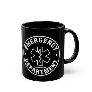 May include: A black ceramic mug featuring a white circular emblem with the text EMERGENCY DEPARTMENT and PARAMEDIC-07. The center of the emblem displays a six-pointed Star of Life symbol with a serpent-entwined staff, all in a clean white print.