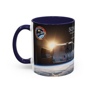 May include: A dark blue ceramic mug featuring a space-themed graphic of the Apollo-Soyuz mission. The design displays a spacecraft orbiting Earth with the sun in the background, the Apollo-Soyuz mission patch, and text reading SOYUZ and USA.