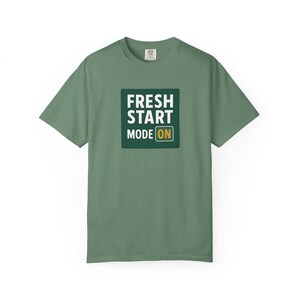May include: Sage green t-shirt with the text "FRESH START MODE ON" in a white font inside a dark green square. The shirt has short sleeves and a crew neck.
