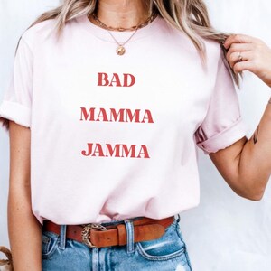 May include: Pale pink t-shirt featuring the text "BAD MAMMA JAMMA" in red. The shirt is worn with blue denim jeans and a brown leather belt.