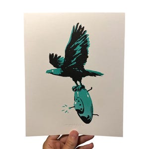May include: A print featuring a black eagle with teal accents, perched atop a teal object with a circular design. The eagle's wings are spread, and the artwork is on a white background. The print has a minimalist, graphic style.