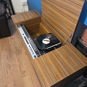 Puede incluir: Un tocadiscos de madera vintage con un panel de control plateado y un disco de vinilo negro en el tocadiscos. El tocadiscos está alojado en un mueble con un patrón de vetas de madera a rayas. La tapa del mueble está abierta.