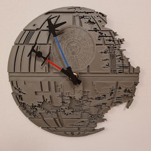 Könnte beinhalten: Graue Death Star Wanduhr mit schwarzen TIE-Fightern und blauen und roten Lichtschwert-Zeigern. Die Uhr hat ein detailliertes, dreidimensionales Design und ist an einer weißen Wand befestigt.