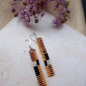 Puede incluir: Pendientes de cuentas hechos a mano con un diseño rectangular. Los pendientes presentan un patrón de bloques de color de cuentas marrones, negras, plateadas y naranjas. Cuelgan de ganchos plateados. Una flor morada está en el fondo.