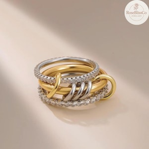 May include: A collection of gold and silver rings. One ring is set with small, clear stones. The other rings are gold and silver. The rings are interlocked. The background is a light beige colour.