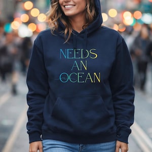 May include: Navy blue hooded sweatshirt with the text "NEEDS AN OCEAN" in a gradient of blue and yellow. The sweatshirt has a front pocket and a drawstring hood. The design is centered on the chest.