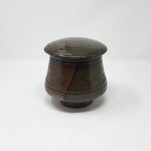 May include: A small, round, ceramic jar with a lid. The jar is brown and green with a speckled glaze.