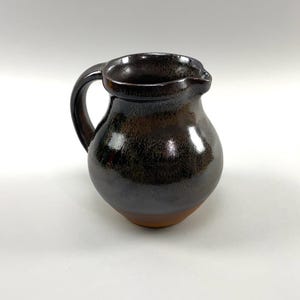 May include: A small, brown ceramic pitcher with a rounded body and a handle. The pitcher has a dark brown glaze with a speckled finish.
