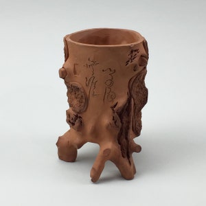 May include: A terracotta-colored ceramic planter shaped like a tree stump. The planter has a rough, textured surface with carved details and Chinese characters. The base has three root-like legs, and the opening is a smooth, round cylinder.