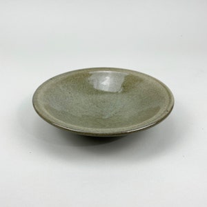May include: A small, round, ceramic bowl with a matte, olive-green finish. The bowl has a shallow depth and a slightly raised rim. The interior features subtle, textured patterns. The bowl is suitable for serving small portions of food or as a decorative piece.