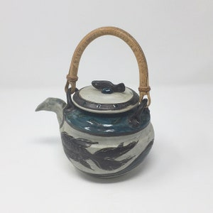 May include: A ceramic teapot with a bamboo handle and a fish fin lid. The teapot is decorated with a blue and white glaze and a black fish design.