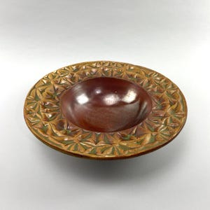 May include: A round ceramic bowl with a smooth, dark reddish-brown center basin. The wide, flat rim features a repeating carved star or leaf pattern with a variegated glaze of earthy brown, tan, and muted green tones throughout the textured surface.
