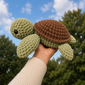 May include: A crochet turtle plush toy featuring a light green head and flippers with a textured brown shell. The turtle has two black plastic safety eyes and is constructed from soft, chunky yarn with visible looped stitches throughout the body.