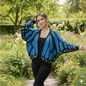 May include: A blue and black butterfly wing-shaped cardigan. The open-front sweater features a vibrant blue wing design with black accents and white dot details. The cardigan is worn over a black top and leggings, set against a backdrop of a garden.