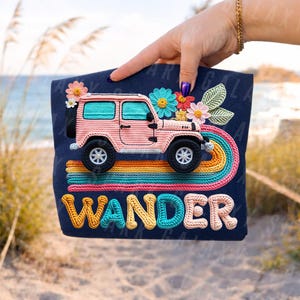 May include: Navy blue tote bag with a pink crochet Jeep, black wheels, a rainbow, and floral details. The word "WANDER" is embroidered in a textured font. The bag is held up by a person.